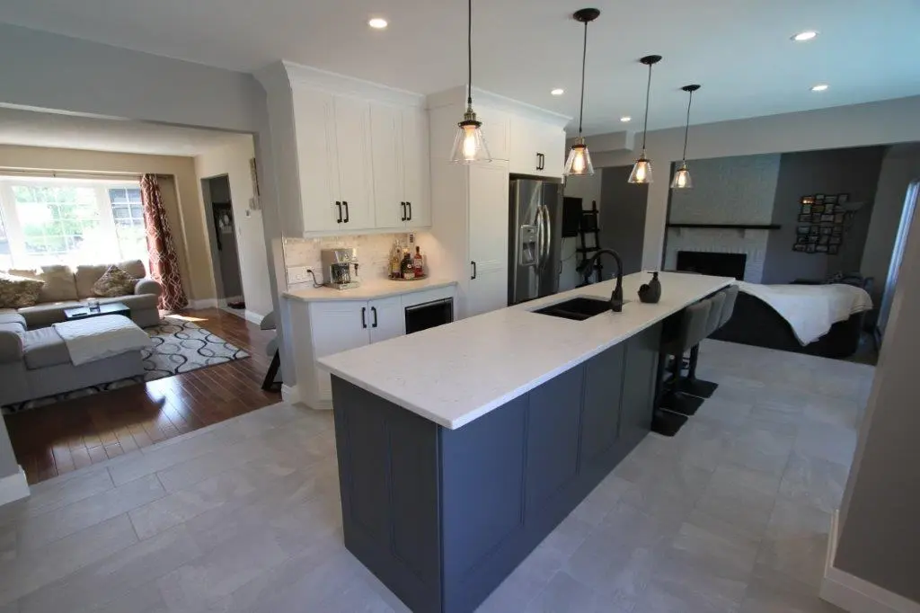 Modern kitchen with a large blue island featuring a white countertop, a black sink, and three pendant lights overhead, adjacent to a living room with a sectional sofa and large window.