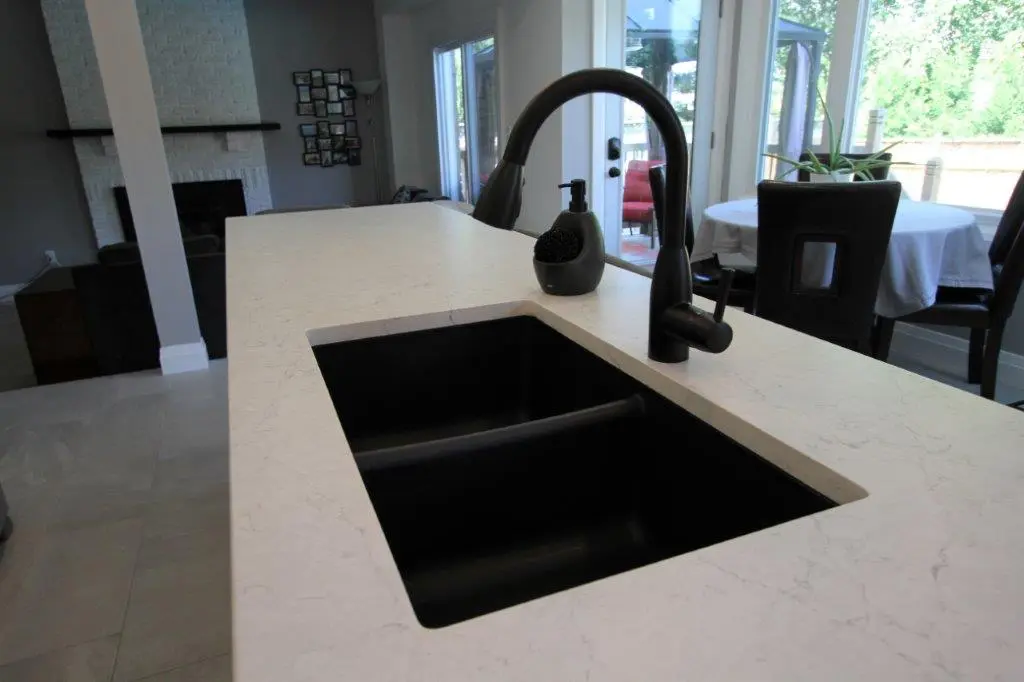 Modern kitchen sink with a black faucet and soap dispenser set in a white quartz countertop, with dining furniture and large windows in the background.