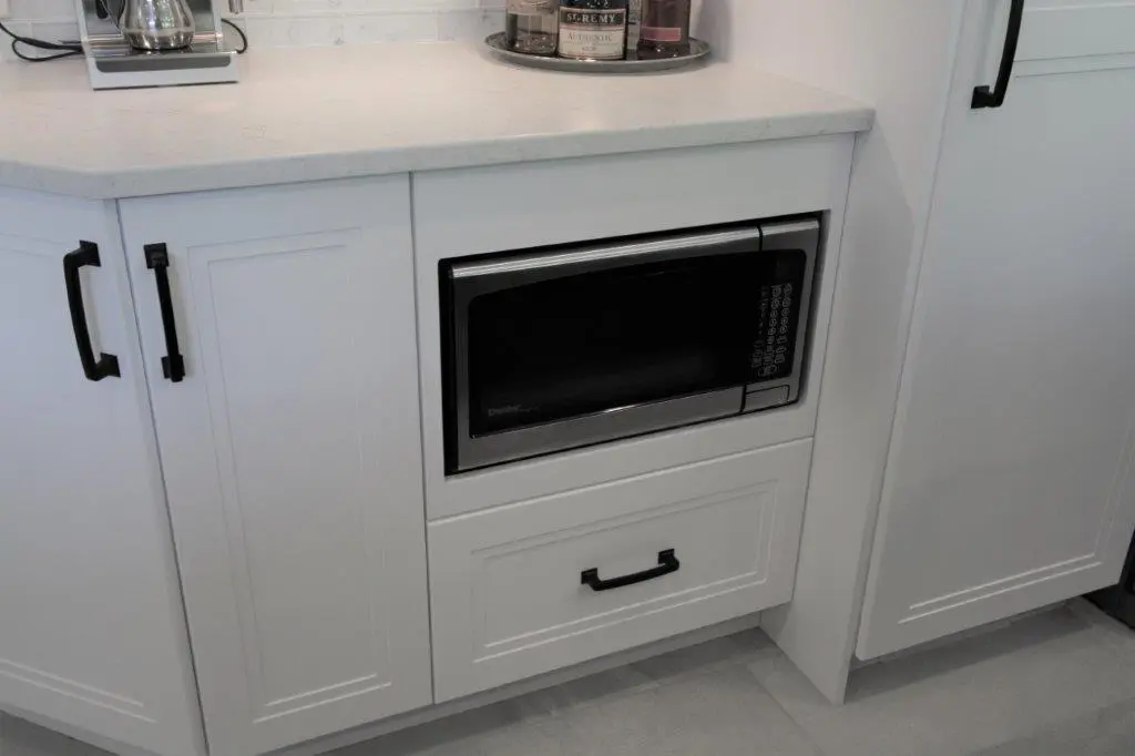 White kitchen cabinetry with black handles featuring a built-in black microwave oven and a large drawer beneath, topped with a light-colored countertop and a round tray holding bottles and jars.