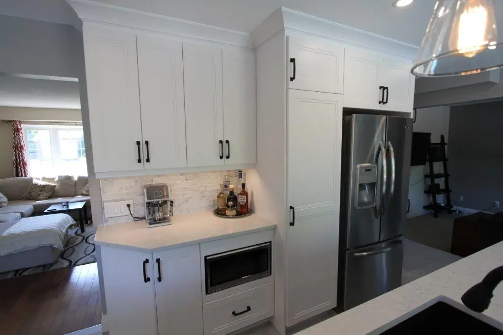 Modern kitchen corner with white cabinets, a built-in microwave, a silver refrigerator, a coffee maker, and bottles on a round tray on the countertop, adjacent to a living room with a large window and beige sofa.