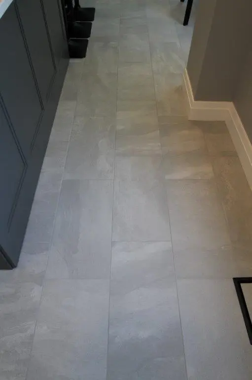 Light gray rectangular floor tiles arranged in a staggered pattern in an indoor setting with a dark cabinet on the left and a beige wall with white baseboard on the right.