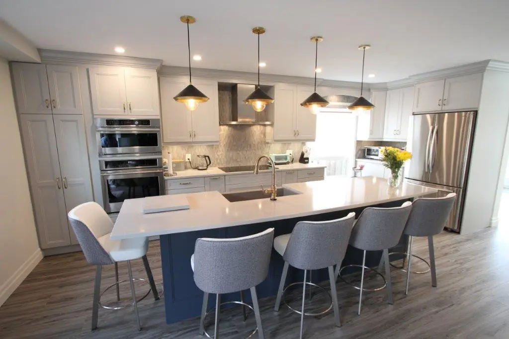 Modern kitchen with a large island featuring a white countertop and five gray upholstered bar stools, surrounded by light gray cabinets, stainless steel appliances, and four black pendant lights hanging above the island.
