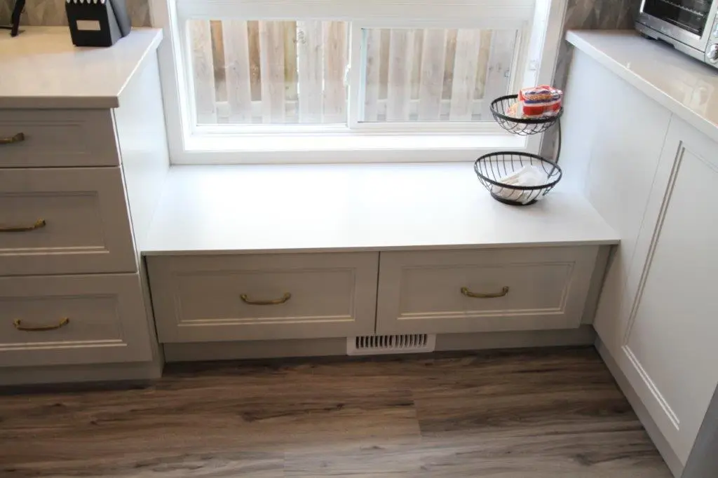 A white window bench with two drawers beneath it, set between two kitchen countertops, featuring brass handles and a wooden floor beneath, with a tiered black wire basket on the bench containing food items.