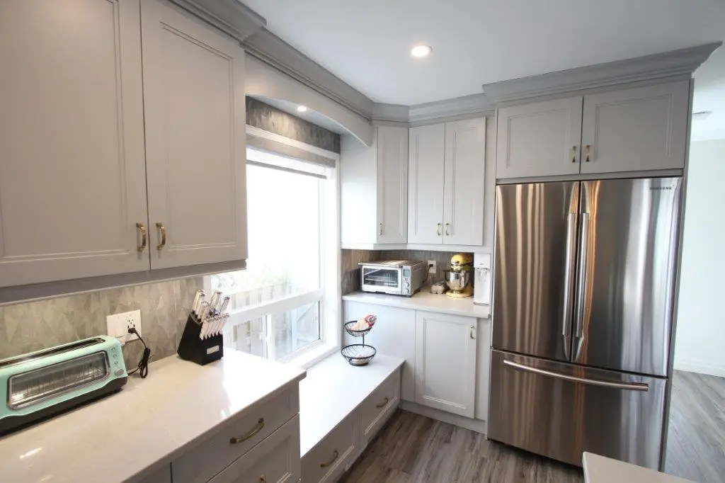 Modern kitchen corner with light gray cabinets, stainless steel French door refrigerator, toaster oven, stand mixer, knife block, mint green toaster, and large window with a built-in bench under it, all on wood flooring.