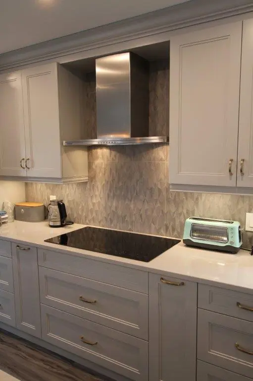 Modern kitchen countertop with light gray cabinets, a built-in black electric cooktop, a stainless steel range hood, a teal toaster, and a coffee maker, set against a textured gray backsplash.