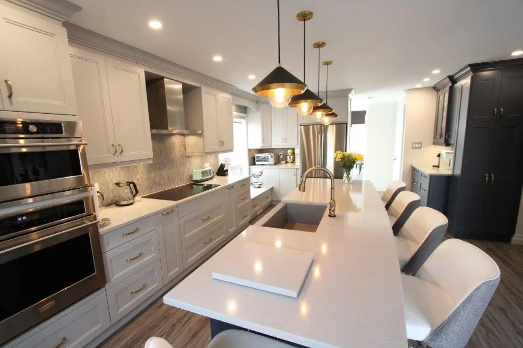 Modern kitchen with white cabinets, stainless steel appliances, a large island with a built-in sink, pendant lights overhead, and several white and gray upholstered chairs along the island.