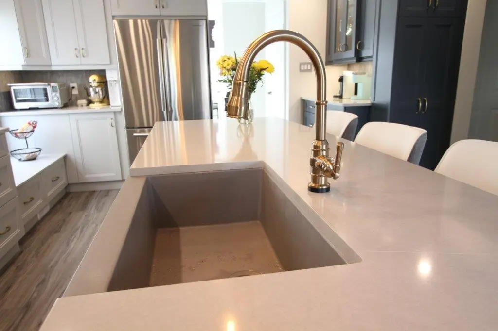 Modern kitchen island with gray countertop, built-in rectangular sink, brass high-arc faucet, white and gray chairs, stainless steel refrigerator, white cabinets, and a vase of yellow flowers in the background.