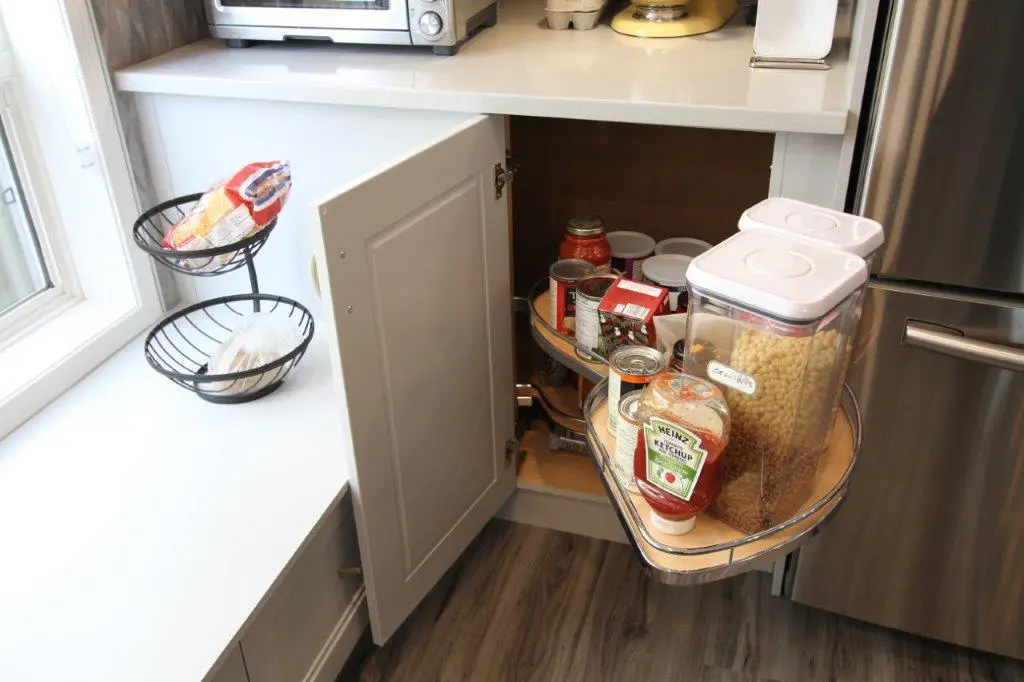 Corner kitchen cabinet with a pull-out shelf containing various canned goods, jars, and containers, next to a stainless steel refrigerator and a countertop with a basket of packaged food items.
