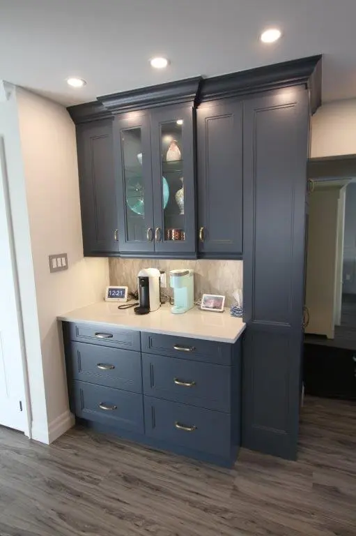 Dark blue built-in kitchen cabinets with glass display doors on top, white countertop holding two coffee makers, a small digital clock, and a framed photo, with hardwood flooring and recessed ceiling lights above
