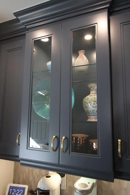 Dark blue kitchen cabinet with glass doors displaying decorative vases and a large turquoise plate inside, below which is a white coffee maker on the counter and a digital clock showing 12:22.