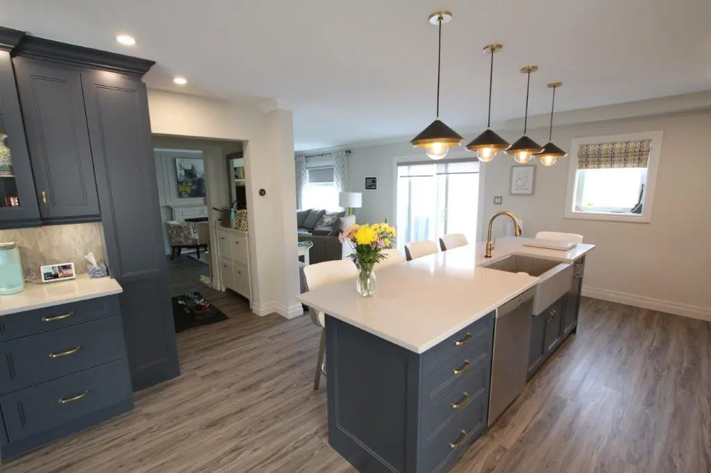 Modern kitchen with a large island featuring a white countertop, dark blue cabinets, a built-in sink with a brass faucet, four pendant lights overhead, and a vase of yellow and pink flowers on the island, with adjacent living room area visible through a doorway and glass sliding doors.