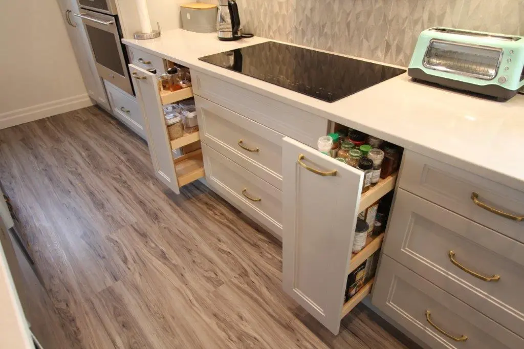 Modern kitchen with light-colored cabinets and wood flooring, featuring two pull-out spice racks filled with spice jars, a black cooktop, and a retro-style turquoise toaster on the white countertop.
