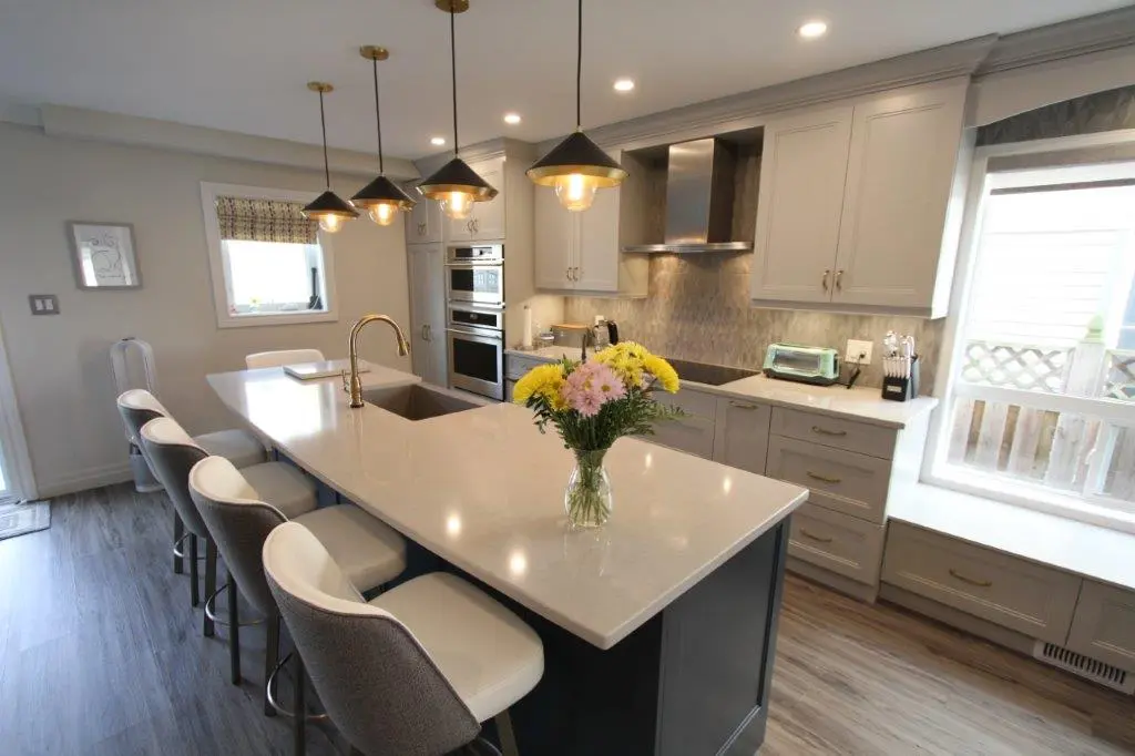 Modern kitchen with a large island featuring a white countertop, gray upholstered bar stools, a vase of mixed yellow and pink flowers, pendant lights, stainless steel appliances, gray cabinets, and a window with a bench seat.