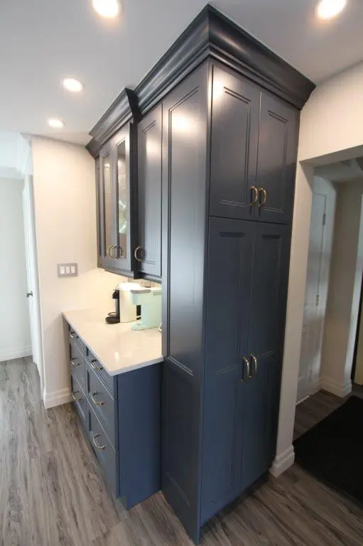 Tall blue kitchen cabinets with gold handles extending to the ceiling, accompanied by a white countertop holding a coffee maker, set against a light-colored wall and wood-look flooring.