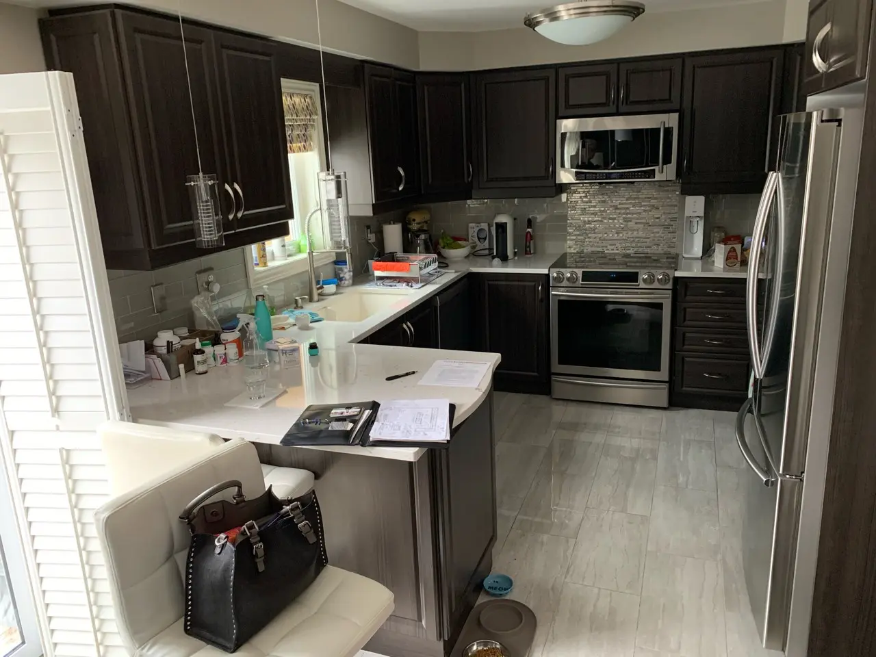 a before picture of a U-shaped kitchen with dark wood cabinets, stainless steel appliances including a refrigerator, oven, and microwave, white countertops cluttered with bottles, a black handbag on a white cushioned bar stool, and pet bowls on the tiled floor near the kitchen island.