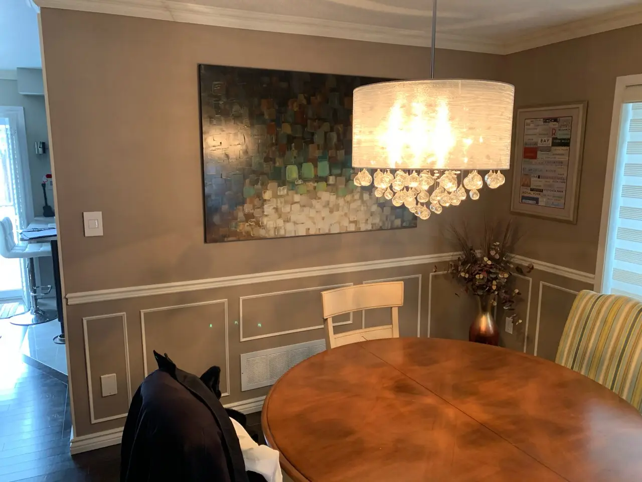 Dining room with a round wooden table surrounded by chairs, a modern chandelier with hanging glass ornaments, abstract wall art, and a floral arrangement in a vase on the corner.