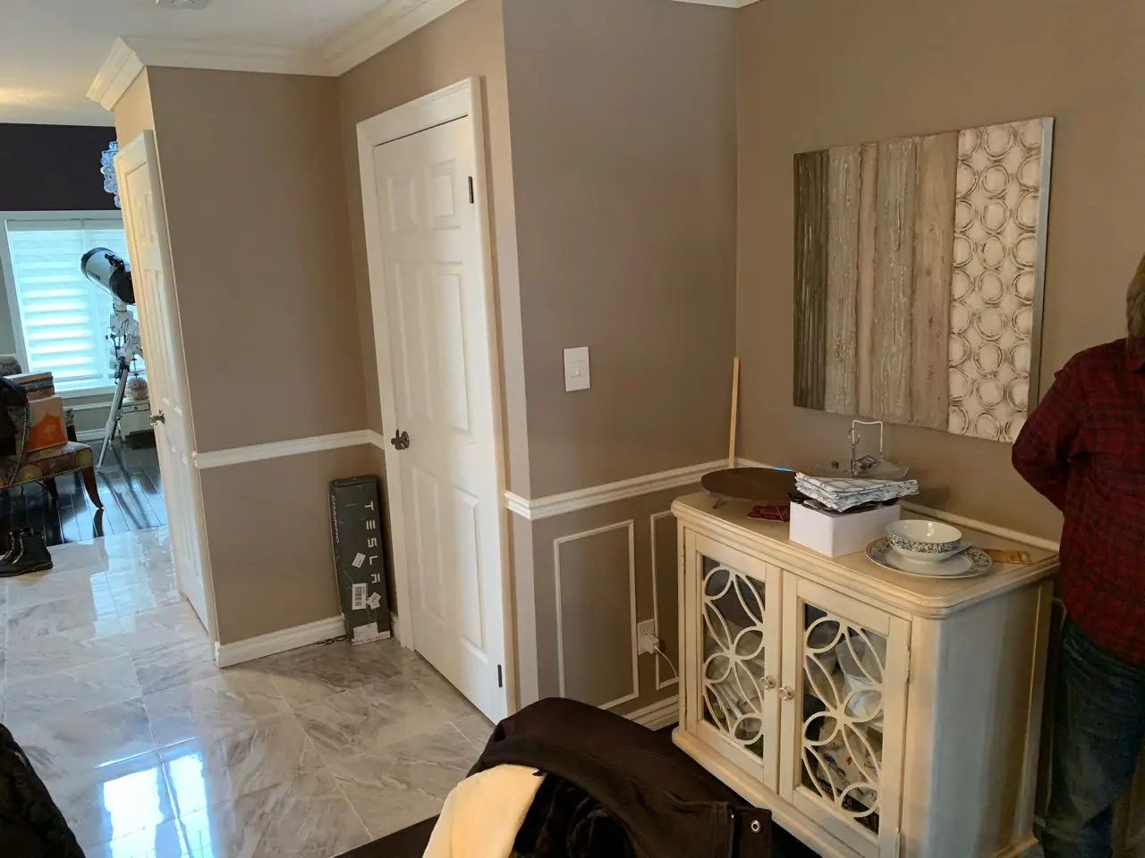 Interior corner of a room with beige walls and marble floor tiles, featuring a white door, a decorative white cabinet with glass-patterned doors topped with dishes and a stack of cloth, a brown and cream wall artwork above, and a person partially visible wearing a red plaid shirt on the right side