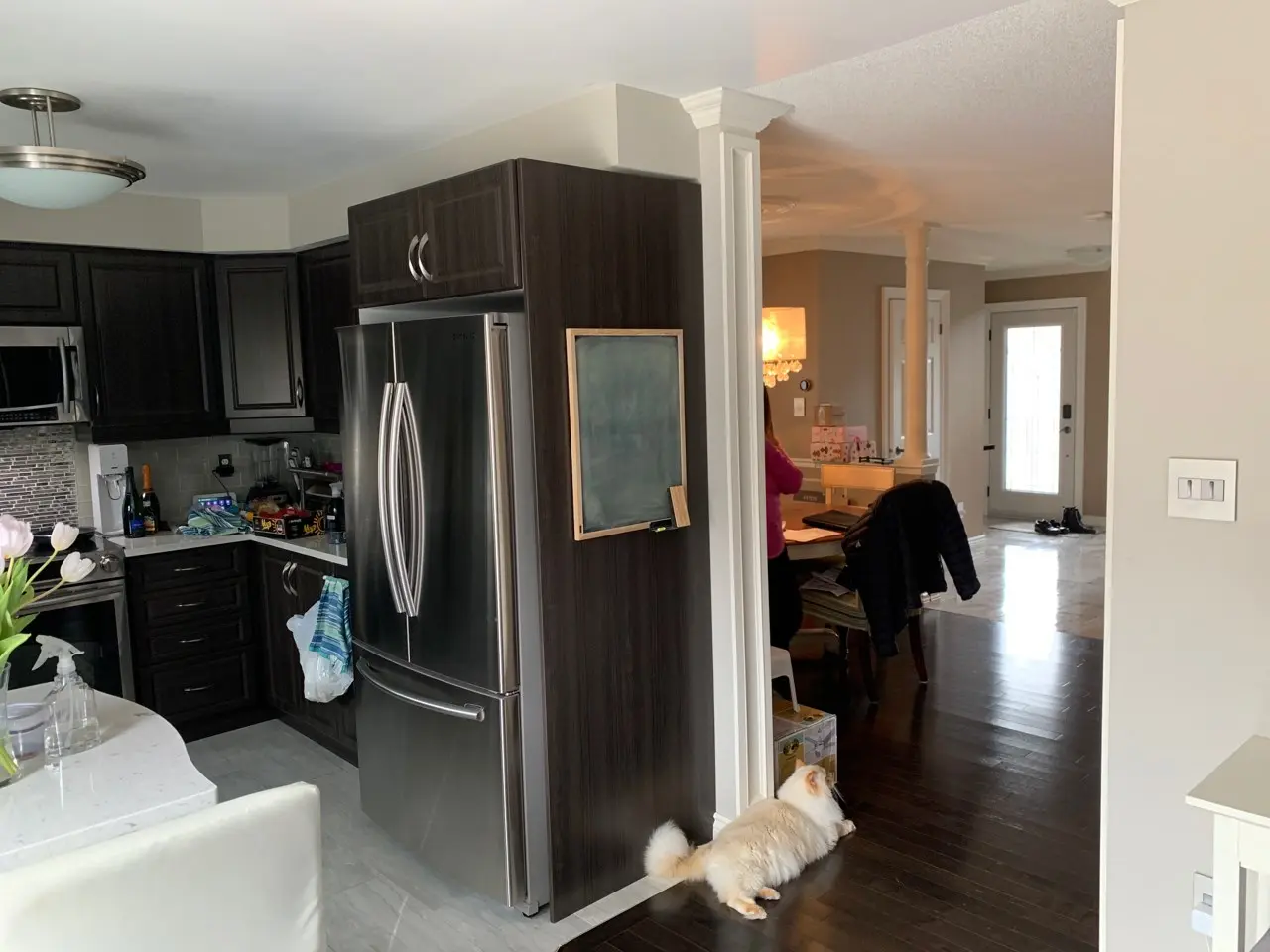 A before renovation Interior view of a kitchen with dark wood cabinets and a stainless steel refrigerator, a white cat lying on a dark wood floor near the entrance to a living area where a person in a pink top is seated at a table with a black jacket on the chair, and sunlight coming through a glass front door in the background.
