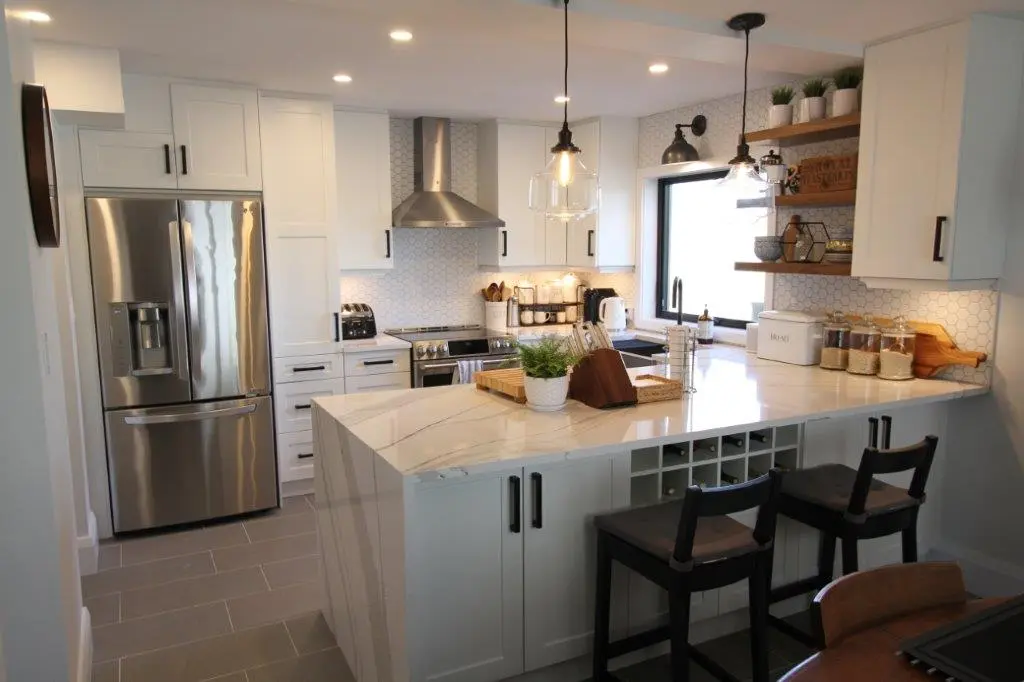 Modern kitchen with white cabinetry, quartz countertop island with two black cushioned chairs, stainless steel refrigerator and stove, hexagonal tile backsplash, pendant and wall lights, and various kitchen accessories on counters and shelves.
