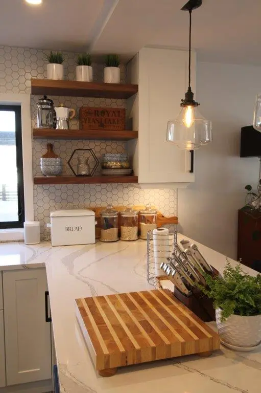 Modern kitchen countertop with a wooden cutting board, a knife holder with knives, a potted plant, glass containers with grains, a white bread box, and floating wooden shelves with decorative items against a white hexagonal tile backsplash.