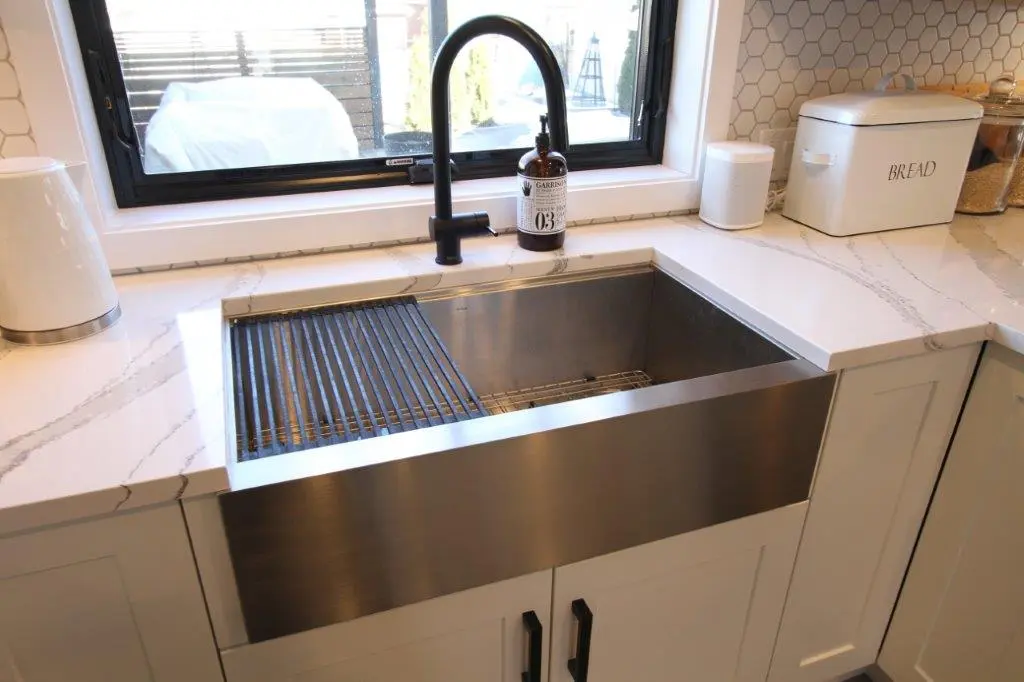Modern kitchen sink area with a large stainless steel apron-front sink, black faucet, soap dispenser, and white quartz countertop, next to a white bread box and countertop appliances.