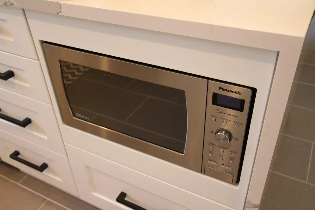 Built-in Panasonic stainless steel microwave oven with digital display and control knob, integrated into white kitchen cabinetry with black handles and gray tiled floor.