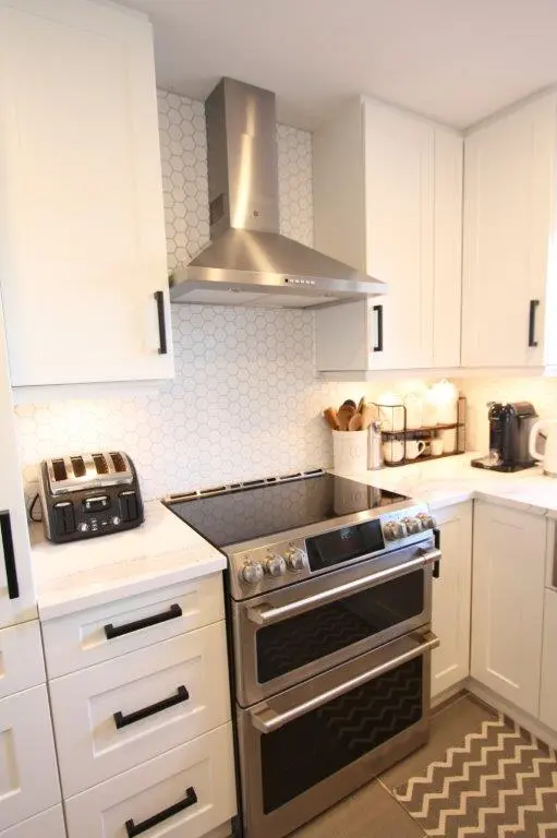 Modern kitchen corner with white cabinets, a stainless steel double oven range with a smooth cooktop, a stainless steel range hood above, a black toaster on the left countertop, and a coffee maker on the right countertop, with a hexagonal tile backsplash and a chevron-patterned rug on the floor.