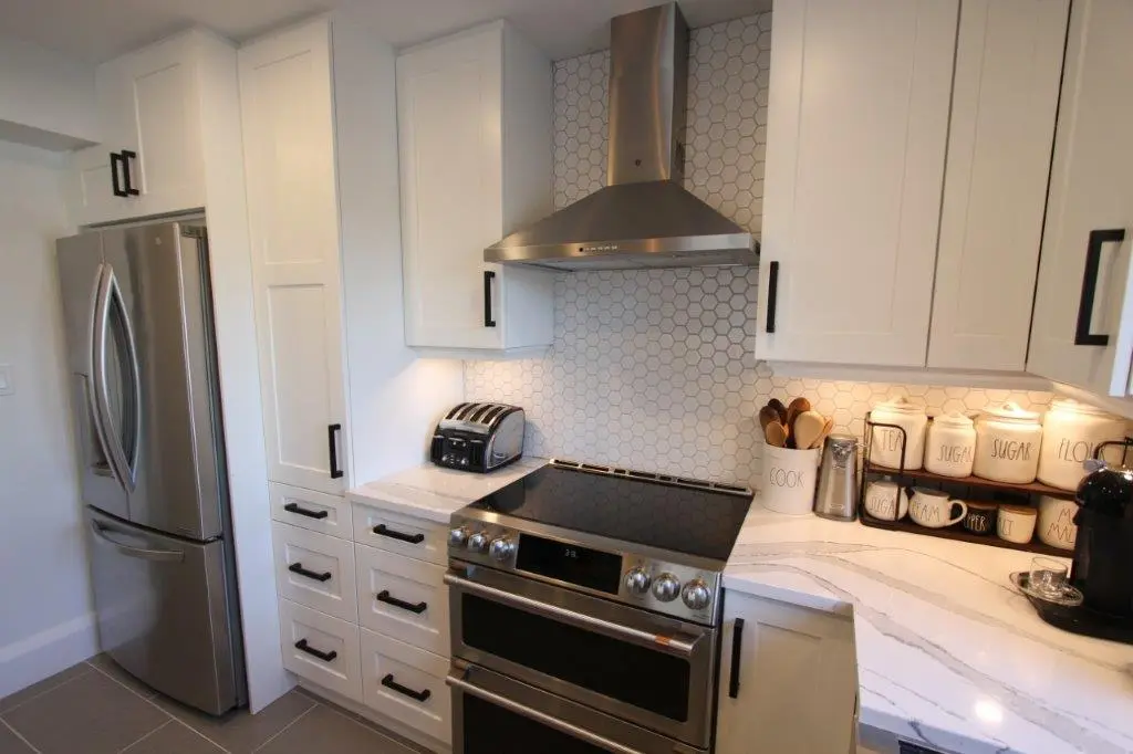 Modern kitchen corner with stainless steel refrigerator and stove, white cabinets with black handles, hexagonal tiled backsplash, and a white quartz countertop holding a toaster, labeled canisters, utensils, and a coffee machine.