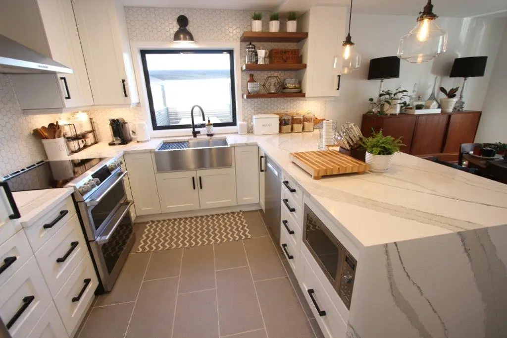 Modern kitchen with white cabinetry, stainless steel appliances including a double oven and dishwasher, a large farmhouse sink, quartz-patterned countertops, wooden shelves with small plants and containers, a wooden cutting board and a potted fern on the counter, hexagonal tile backsplash, and pendant lights above the countertop extending into a dining area with plants and dark wood furniture.