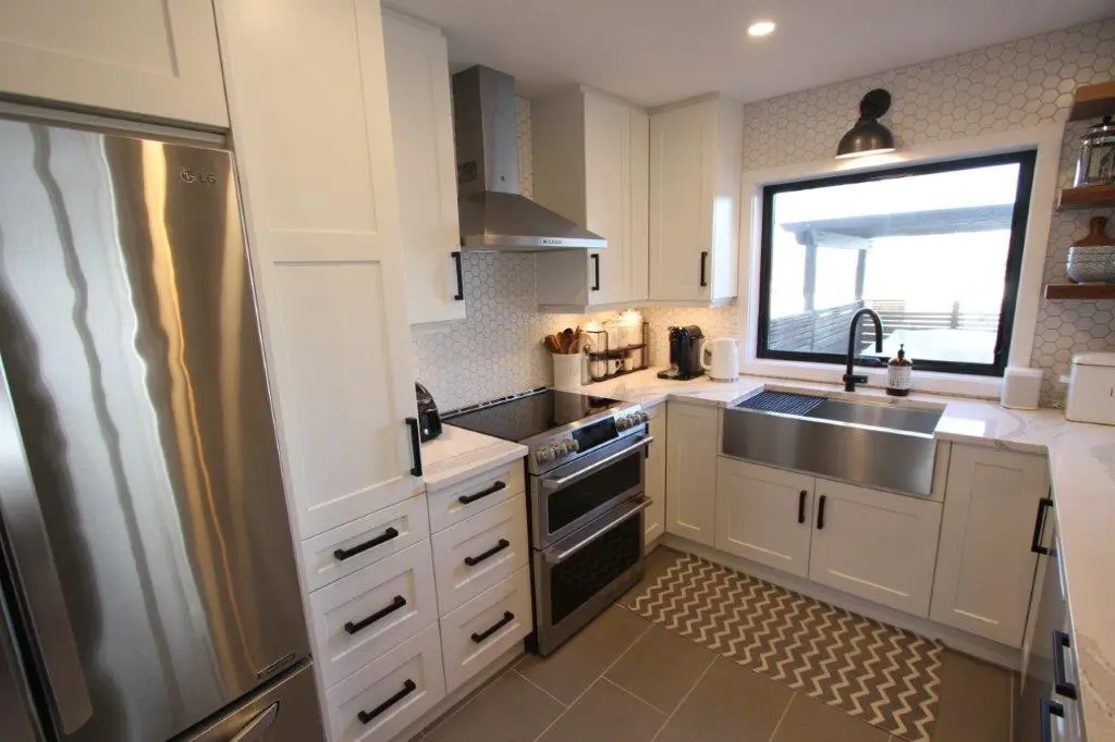 Modern kitchen with white cabinets, stainless steel appliances including an LG refrigerator and a double oven, a large stainless steel farmhouse sink under a window, hexagonal tile backsplash, and a zigzag patterned rug on the gray tiled floor.