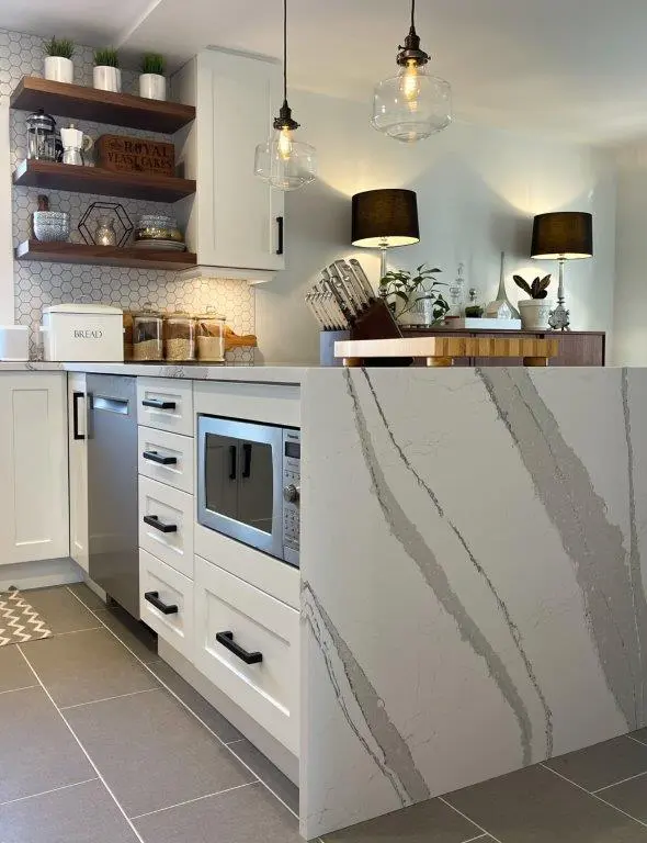 Modern kitchen corner featuring white cabinets with black handles, stainless steel dishwasher and microwave, quartz countertop with gray veins, wooden shelves with potted plants and decorative items, and hanging glass pendant lights over the counter.