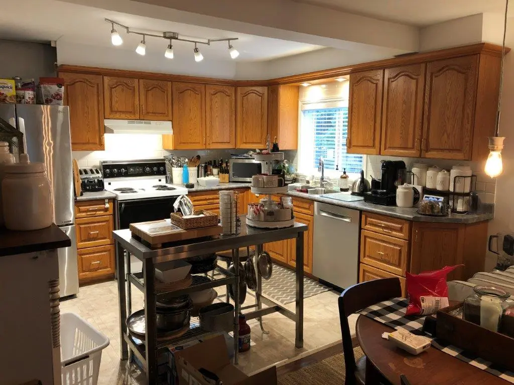 A before pic of a Kitchen with wooden cabinets, a white stove and range hood, a stainless steel refrigerator, a central metal island with kitchen items, and a dining table with a black and white checkered cloth and various items on it.