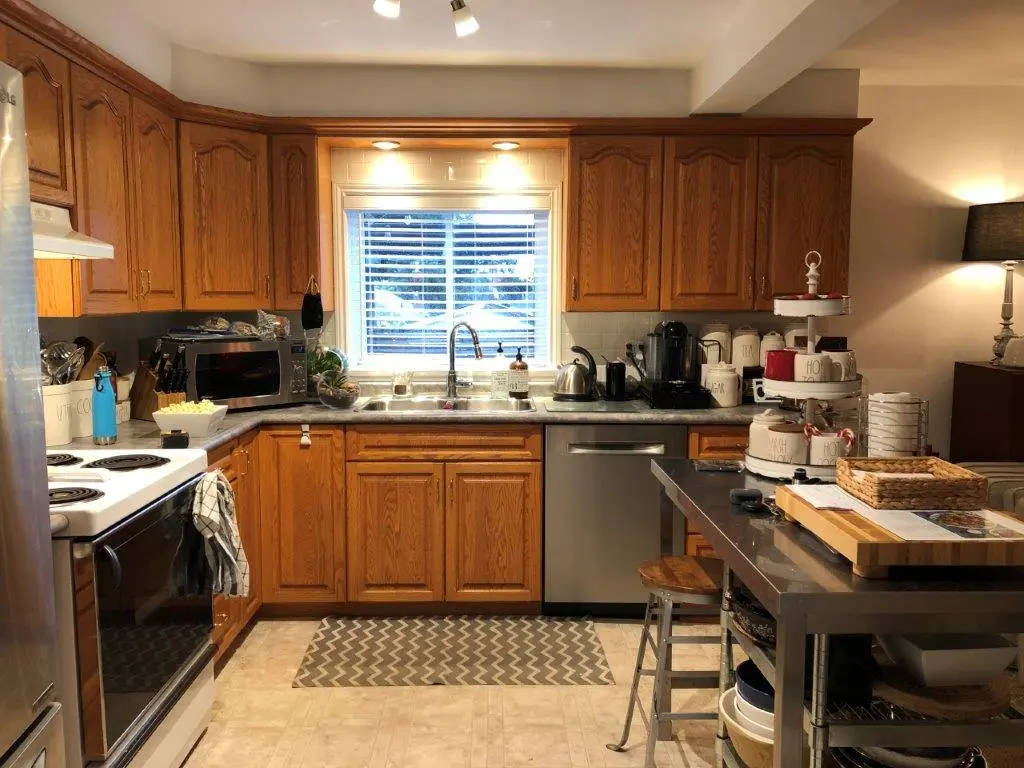A before pic of a kitchen with wooden cabinets, a window above the sink with blinds, stainless steel dishwasher, white electric stove, microwave, and a metal kitchen island with various containers and a three-tiered tray on top.