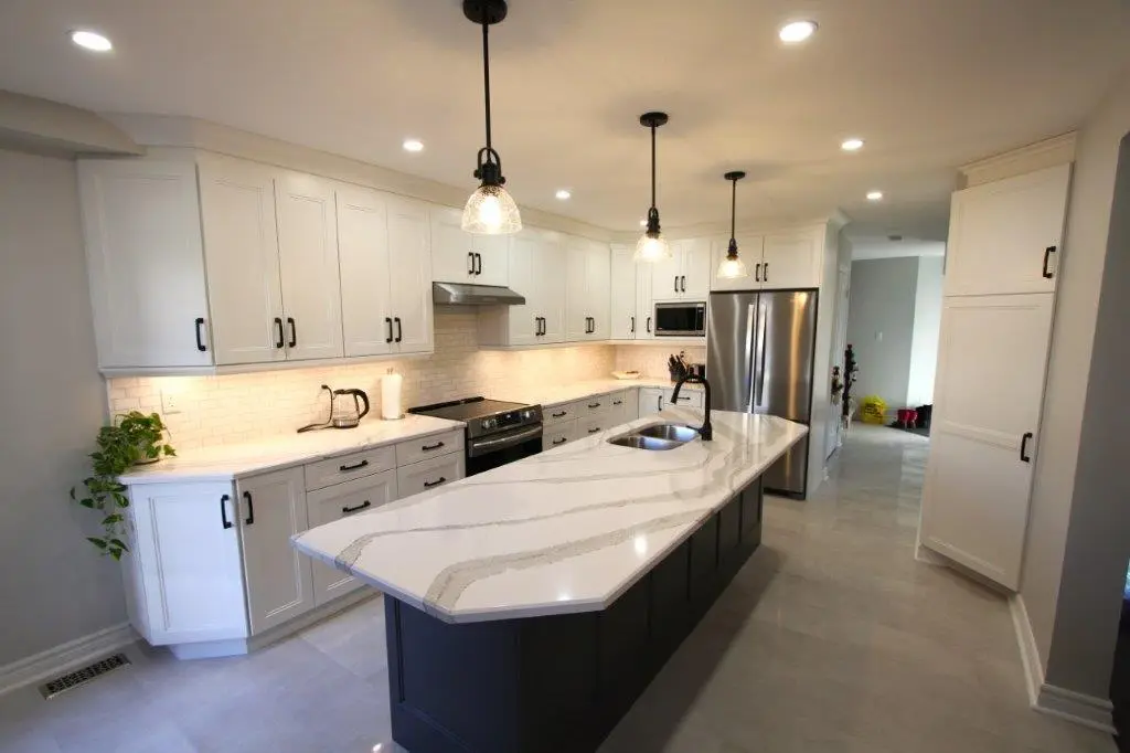 Modern kitchen with white upper and lower cabinets, stainless steel appliances, a large quartz kitchen island with a built-in sink and black base, pendant lighting, and light gray flooring.