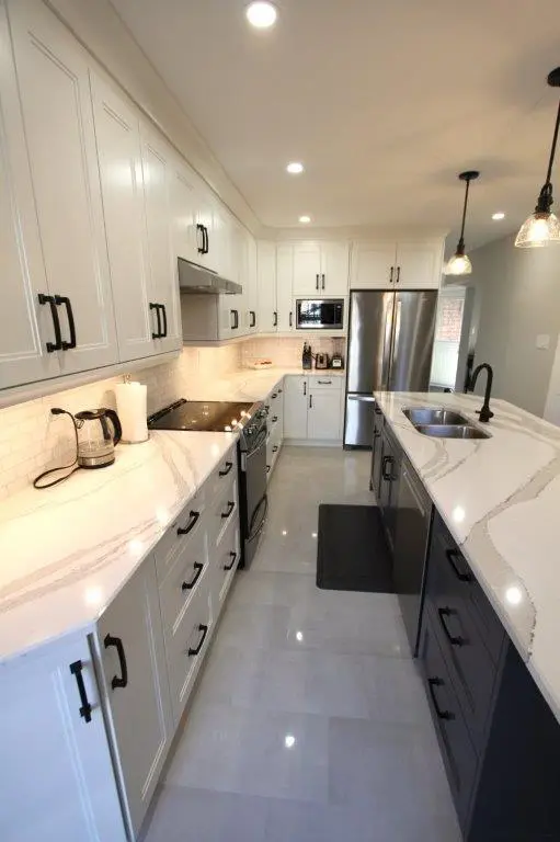 Modern kitchen with white upper and lower cabinets featuring black handles, quartz countertops, a stainless steel stove and refrigerator, a sink with a black faucet on a dark island, and pendant lights hanging from the ceiling.