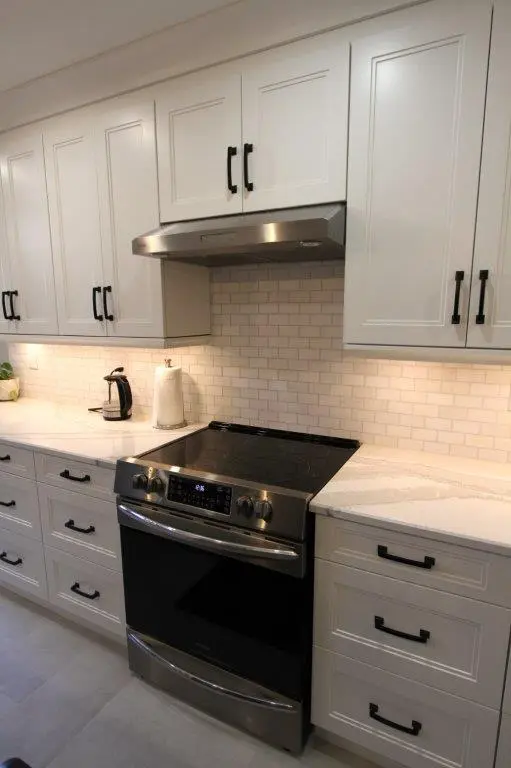 Modern kitchen with white cabinets, black handles, a stainless steel electric stove with digital controls, quartz countertops, white subway tile backsplash, a stainless steel range hood, a black electric kettle, and a paper towel holder under cabinet lighting.