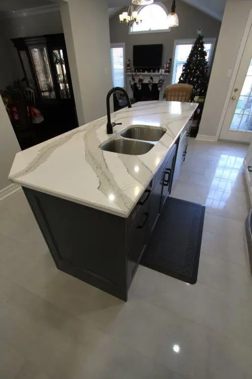 Modern kitchen island with white quartz countertop and built-in double sink, black faucet, dark cabinetry, and a black floor mat in front of it, with a living room in the background featuring a Christmas tree, a fireplace with stockings, and a TV.