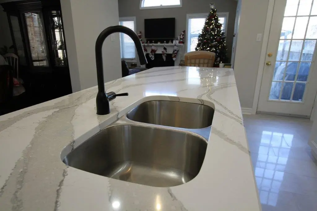 Modern kitchen with a white quartz countertop featuring a double stainless steel sink and a black faucet, with a decorated Christmas tree, stockings on a fireplace mantel, a TV, and a large windowed door in the background.