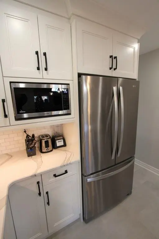 Stainless steel French door refrigerator with bottom freezer in a modern kitchen, surrounded by white cabinetry with black handles, and appliances including a microwave, toaster, and blender on a white quartz countertop with a subway tile backsplash.