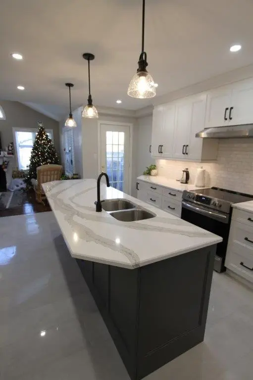 Modern kitchen with a large gray island featuring a white quartz countertop and dual sinks, three hanging pendant lights above, white cabinets with black handles, a stainless steel oven, and a decorated Christmas tree in the background near a window and rocking chair.