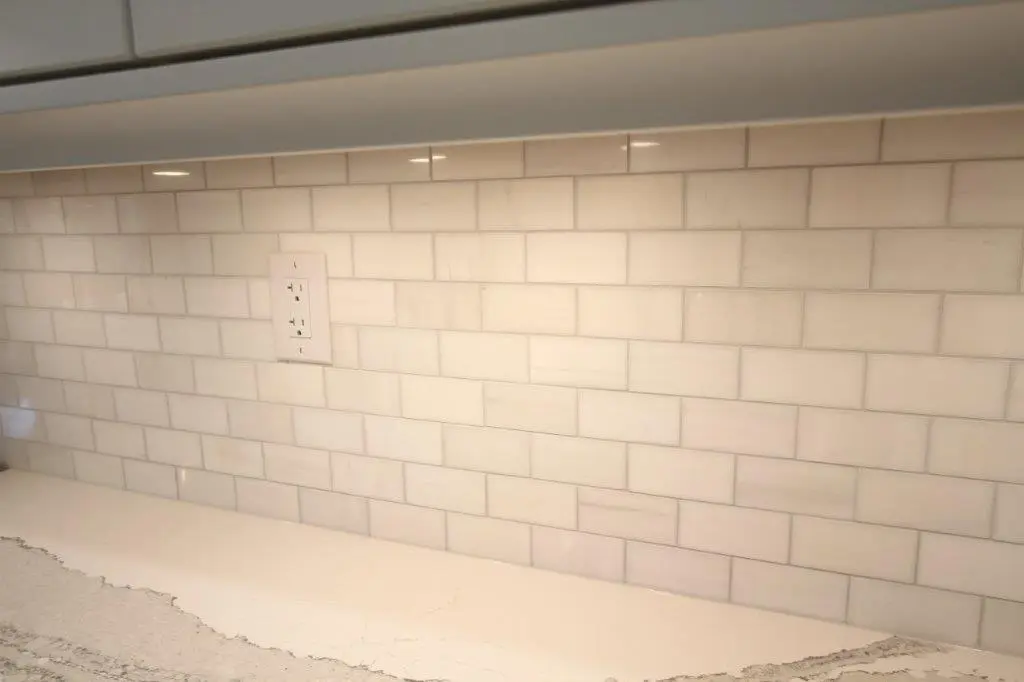 Kitchen backsplash composed of light-colored subway tiles with a white electrical outlet in the middle and a light-colored countertop below.