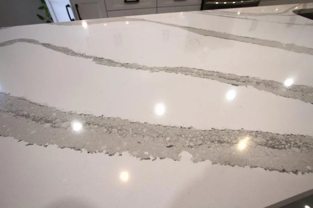 Close-up of a polished white quartz kitchen countertop with wide gray veining, reflecting ceiling lights and showing part of a kitchen cabinet in the background