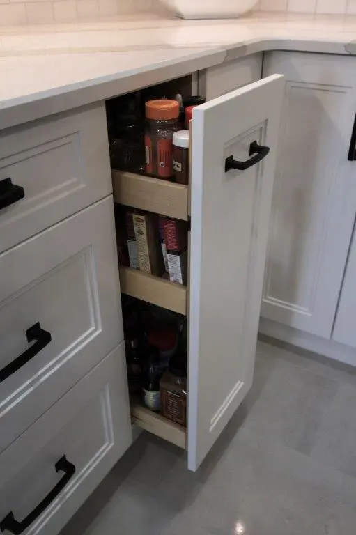 Open white kitchen cabinet with black handles revealing three wooden pull-out shelves stocked with various spice jars and seasoning containers.