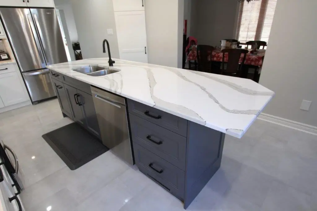 Modern kitchen island with a white quartz countertop featuring gray veining, equipped with a double stainless steel sink and a black faucet, dark gray cabinetry with black handles, and a built-in stainless steel dishwasher, set in a kitchen with light gray tile flooring and stainless steel refrigerator in the background.