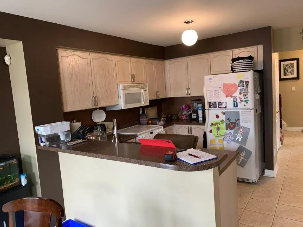 a before picture of a kitchen with dark brown walls, cream-colored cabinets, a white refrigerator covered with children's artwork, a countertop with various items including a red cutting board and a notebook, and a fish tank on the left side.