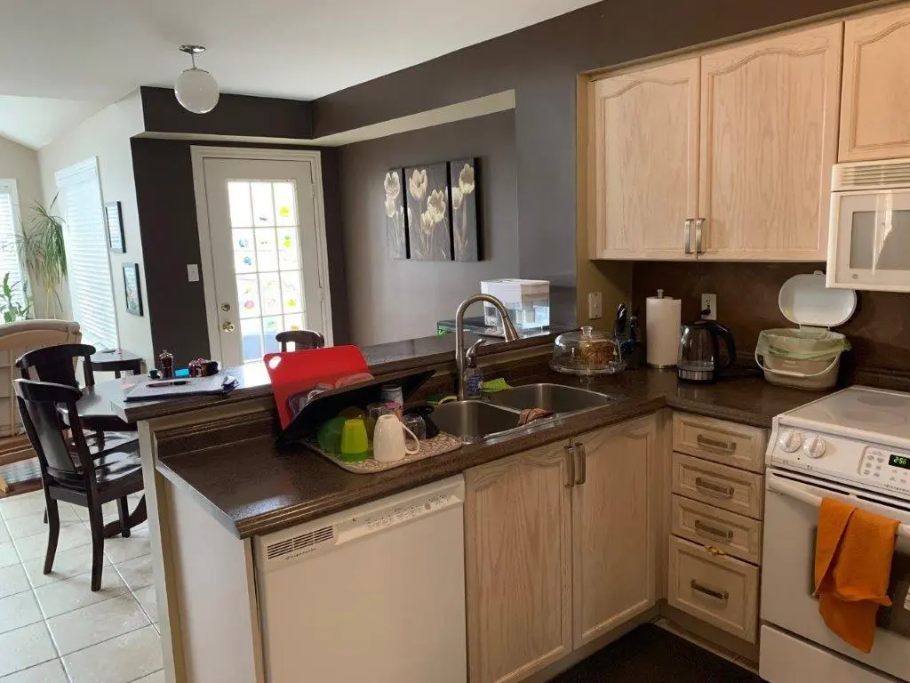 A before picture of a Kitchen interior with light wood cabinets, a dual sink with dishes, a white stove with an orange towel, a microwave above, and a dining area with dark wooden chairs and a glass door in the background.