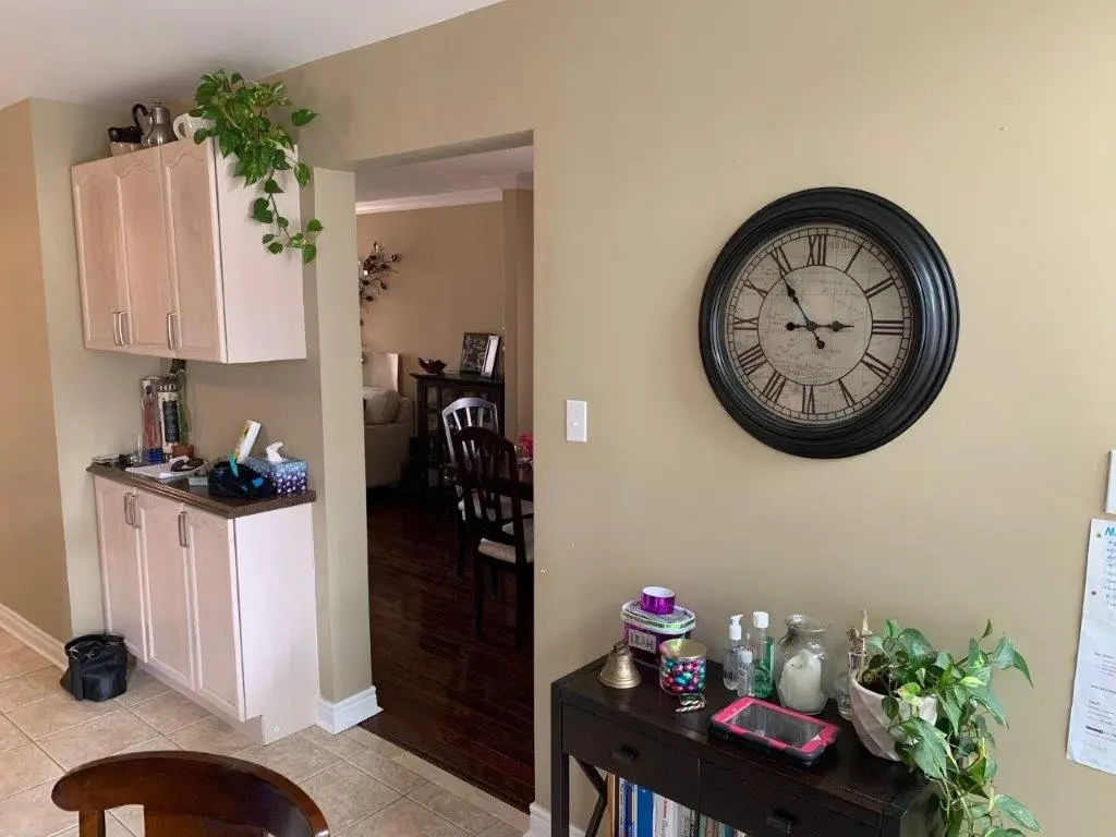 A before picture of A beige room corner with light wood cabinets on the left wall and a black console table on the right wall underneath a large round wall clock with Roman numerals. The console table holds various items including plants, containers, hand sanitizers, a tablet, and a small decorative bell. The floor is tiled, and a doorway in the middle leads to a room with dark wood floors and wooden dining chairs visible.