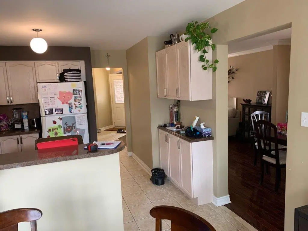 A before View of a kitchen and dining area with beige walls and tiled and wood floors, featuring a white refrigerator with magnets and drawings, light-colored cabinets, a small countertop with various items, a plant hanging from the upper cabinets, and dark wooden chairs around a glass dining table in the adjacent room.