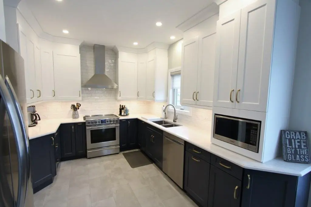 Modern U-shaped kitchen with white upper cabinets, dark lower cabinets, stainless steel appliances including refrigerator, stove with hood, dishwasher, and microwave, white countertops, subway tile backsplash, and a small window above the double sink.
