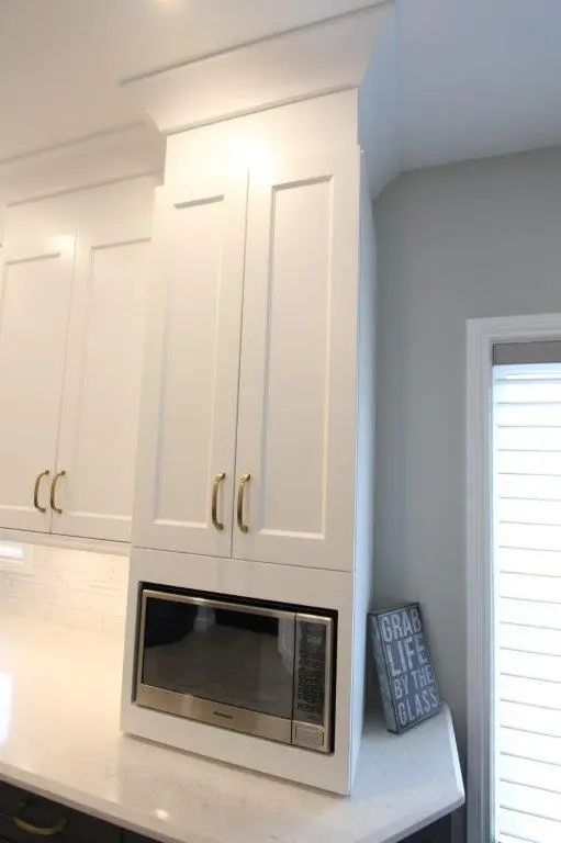 White kitchen cabinets with brass handles, a built-in microwave below, and a countertop with a sign that reads 'Grab life by the glass' next to a window with blinds.